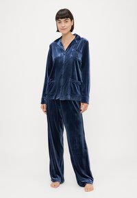 Conjunto de pijama em veludo azul marinho, com uma parte superior de botão e dois bolsos frontais, e calças largas combinando. Textura suave, design clássico.