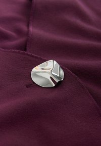 Sølvbroche med et buet, abstrakt design, placeret på et tekstureret mørk bordeauxstof. Brochen har en poleret overflade og diskrete konturer.