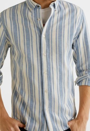 Mężczyzna ubrany w jasnoniebieską i białą koszulę w pionowe paski, z podwiniętymi rękawami i rękami w kieszeniach.