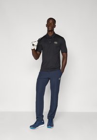 Polo noir avec le logo EAZ, pantalons bleu marine et chaussures de sport bleues. Tenant un club de golf avec un gant blanc sur une main.