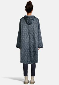 Waterdichte poncho in marineblauw met een geometrisch bladmotief, voorzien van een capuchon en wijde mouwen, aangevuld met strakke donkere broeken en lichte laarzen.