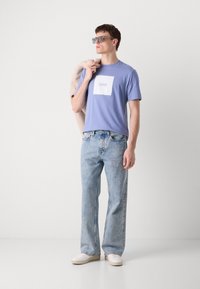 T-shirt de algodão lilás claro com um patch de logo em forma de quadrado, combinado com jeans de lavagem clara de cintura alta e sapatos brancos. Modelo segurando uma jaqueta bege.