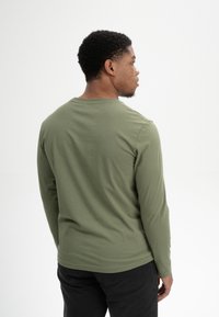 Langarmhemd in olivgrün aus weichem Stoff, mit klassischem Rundhalsausschnitt und glatter Textur, getragen mit schwarzen Hosen.