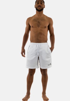Homme torse nu debout pieds nus portant un short blanc à cordon avec poches et logo « Sergio Tacchini » sur la jambe gauche.