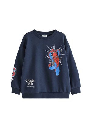 Barn iført marineblå Spider-Man sweatshirt og bukser, blå strikket hat samt røde og hvide sneakers, smilende og stående mod en ensfarvet baggrund.