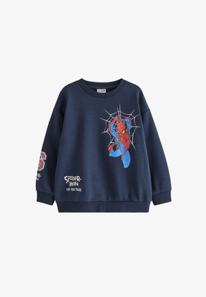 Kind draagt donkerblauwe Spider-Man trui en broek, blauwe gebreide muts en rode en witte sneakers, glimlachend en staand tegen een effen achtergrond.