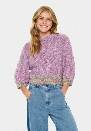 Blonde Frau lächelt, trägt einen lila und beigen gestrickten Pullover mit Ballonärmeln und blaue High-Waist-Jeans mit Händen in den Taschen.