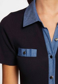 Nära bild av en svart ribbad kortärmad topp med krage i blå denim, knappslå och en liten bröstficka med metallsnapknappar.