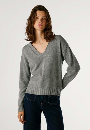 Grauer V-Ausschnitt-Pullover mit gerippten Bündchen und Saum, aus weichem Material gefertigt, im entspannten Schnitt mit tiefem Ausschnitt. Kombiniert mit dunklen Jeans.