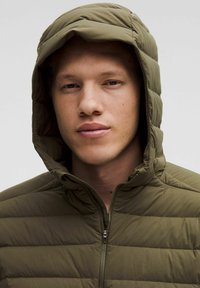 Giacca puffer verde oliva con cappuccio, caratterizzata da un design trapuntato e chiusura con zip. Il tessuto strutturato conferisce un aspetto morbido e isolato.