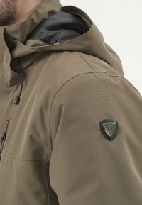 Brun vattentålig jacka med svart innerfoder, justerbar huva och en logotypapplikation på axeln. Slätt strukturerad tyg.