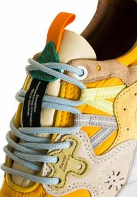 Sneaker giallo realizzato con materiali misti, con lacci blu e grigi, un'etichetta di accento verde e camoscio testurizzato sulla linguetta e sui pannelli laterali.