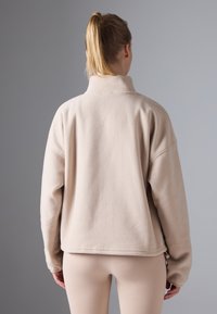 Pulover din fleece bej cu guler înalt, croială lejeră și mâneci lungi. Textură netedă cu un finisaj curat. Fundal gri deschis.