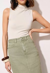 Lichtbeige mouwloze top met hoge kraag, gecombineerd met een olijfgroene denim rok met hoge taille. Het model draagt meerdere bengelarmbanden om één arm.
