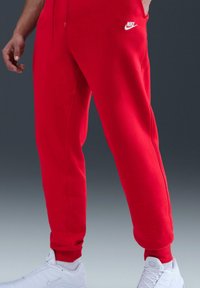 Rode fleece joggingbroek met een aansluitend ontwerp, elastische boorden en een wit Nike-logo op de linker dij. Draagt witte sneakers.