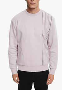 Sudadera de color púrpura claro con mangas largas y puños acanalados. Presenta líneas abstractas negras impresas en la parte frontal, con un diseño de corte relajado.