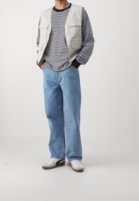 Person som bär en ljusbeige arbetsväst över en svart och vit randig långärmad skjorta, ljusblå jeans och vita sneakers med svarta detaljer.