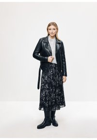 Czarna skórzana kurtka motocyklowa na jasnoszarym, dzianinowym sweterze, w połączeniu z plisowaną czarną spódnicą maxi w kwiaty i czarnymi, sięgającymi do kolan botkami.