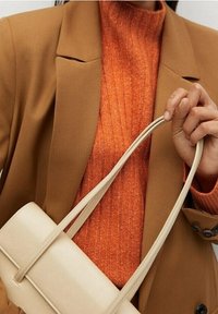 Sac à main en cuir beige avec de longues sangles pour l'épaule tenu d'une main, associé à un pull côtelé orange et un blazer marron.