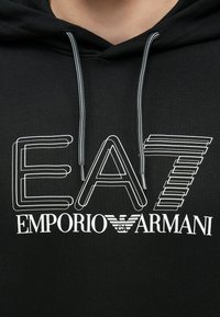 Čierna mikina s kapucňou s výraznou bielou grafikou s nápisom "EA7 EMPORIO ARMANI". Šnúrky v kontrastnej farbe. Mäkká textúra, štandardný strih.