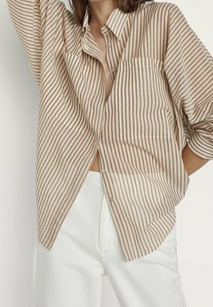 Femme portant une chemise oversize beige et blanche à rayures verticales avec poche avant et pantalon blanc, posant avec le bras levé.
