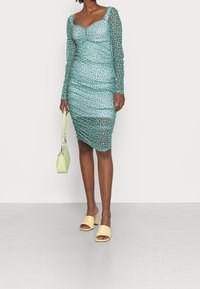 Robe en maille teal à manches longues, design froncé, pois blancs, associée à un sac à main vert et des talons ouverts jaunes.