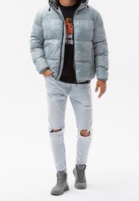 Hellblauer Pufferjacke mit Kapuze, die eine gesteppten Textur aufweist. Darunter ein schwarzes Grafik-T-Shirt, kombiniert mit zerrissenen hellen Jeans und grauen Stiefeln.