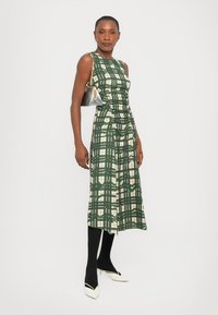 Rochie cu imprimeu tartan verde și crem, cu un corset ajustat, detalii plisate și lungime midi. Asortată cu colanți negri și pantofi albi cu toc, ținând în mână o geantă argintie.