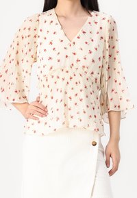 Mujer que lleva una blusa de crepé floral color crema con mangas acampanadas y una falda blanca texturizada con un detalle de botón marrón.