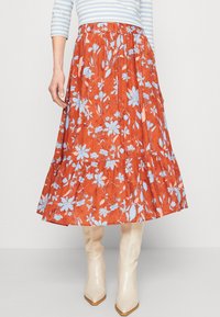 Gemusterter Rock aus orangefarbenem Stoff mit blauen Blumen, ausgestattet mit einem elastischen Taillenbund und einem ausgestellten Saum. Kombiniert mit beigen kniehohen Stiefeln.