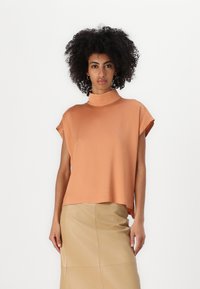 Kortermet topp med høy hals i lys oransje, laget av glatt stoff, parret med et beige skjørt til kneet, med en strømlinjeformet og tettsittende design.