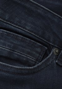 Ciemnoniebieskie dżinsy z bliskim ujęciem przednich kieszeni, widocznym szwem i metalowym guzikiem, ukazujące fakturę i materiał.