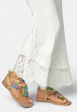 Pies con sandalias beige decoradas con flores de cuentas de colores, combinadas con pantalones blancos que tienen un borde de encaje floral en el dobladillo.