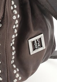 Bolso de cuero marrón con tachuelas ovaladas plateadas a lo largo de un lado y una etiqueta metálica rectangular que muestra "AS 98". Textura suave, diseño blando.