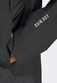 Juodas nepralaidus vandeniui striukė su glotnia tekstūra, ant rankovės yra užrašas „RAIN.RDY“ baltos spalvos, ir šone yra kišenė rankoms.