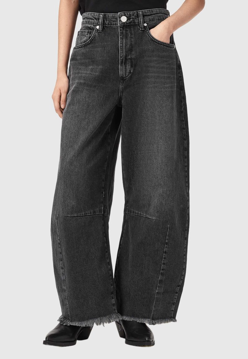 Personne portant un jean denim noir taille haute à jambes larges avec des ourlets effilochés et des bottes noires à talons, une main dans la poche, sur un fond uni.