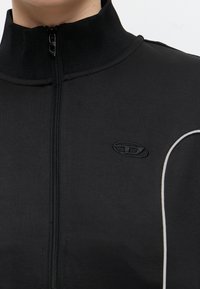 Veste noire zippée avec un col montant, avec un petit logo ovale brodé et un liseré blanc sur l’épaule.