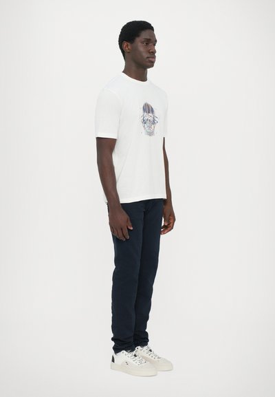 T-shirt blanc à manches courtes avec un imprimé graphique coloré sur le devant, associé à un pantalon bleu marine et des baskets blanches avec des accents noirs.
