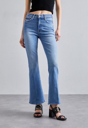 Donna che indossa jeans a zampa a vita alta di colore azzurro chiaro e sandali neri con tacco e dettagli con fibbia dorata, in piedi su un pavimento grigio.