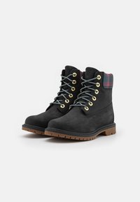 Botas de tobillo de ante negro con ojales dorados, cordones grises y un acento de cuadros en el cuello. Suela de goma resistente para mayor tracción.