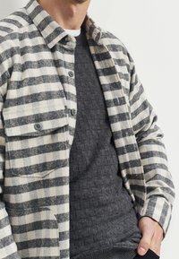 Uldskjortejakke med et sort og hvidt ternet mønster, der har to brystlommer og en grå tekstureret sweater under.