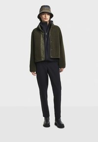 Kurtka z polaru w oliwkowym kolorze z pikowaną oliwkową podszewką, czarną warstwą wewnętrzną oraz czarne dopasowane spodnie, zestawione z beżowym kapeluszem typu bucket i czarnymi butami.