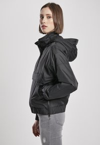 Črna zip-up jakna s kapuco, ki ima sprednji žep, elastične manšete in krojeno kroj. Model nosi svetlo siva raztrgana kavbojka.