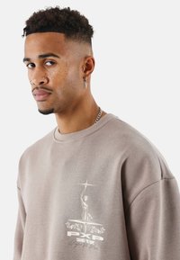 Sudadera marrón claro con cuello redondo, que presenta un diseño gráfico en blanco en el pecho y una tela texturizada.