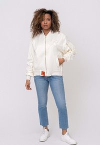 Crème bomberjack met een ritssluiting aan de voorkant, geribde manchetten en een geborduurd logo. Gecombineerd met lichtblauwe recht gesneden jeans en witte sneakers.