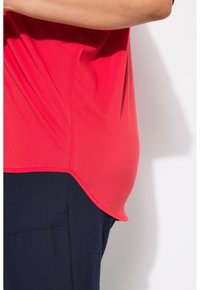 Top rosso a maniche corte con orlo curvo e texture liscia, abbinato a pantaloni blu scuro. Focusa sul design fluido del top e sul contrasto di colore.