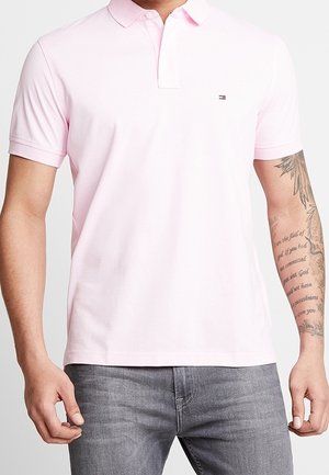 Homme portant un polo rose clair à manches courtes avec un petit logo sur la poitrine et un jean gris, tatouage visible sur l'avant-bras droit.