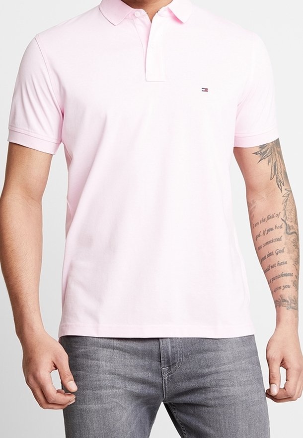 Homme portant un polo rose clair à manches courtes avec un petit logo sur la poitrine et un jean gris, tatouage visible sur l'avant-bras droit.