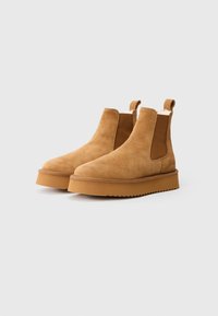 Bruine suède enkelboots met elastische zijkanten, treklipjes en een platte, getextureerde rubberen zool. Rond neustype met naadloze stiksels.