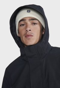 Zwarte waterdichte jas met een hoge kraag, voorzien van een verborgen capuchon. Het model draagt een crèmekleurige geribbelde beanie met een klein logo-opdruk.